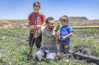 Syrian father, kneeling, and sons engage with each other in an agricultural setting.