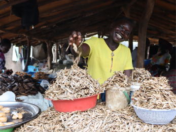 Ugandan woman stands in her shop smiling.
